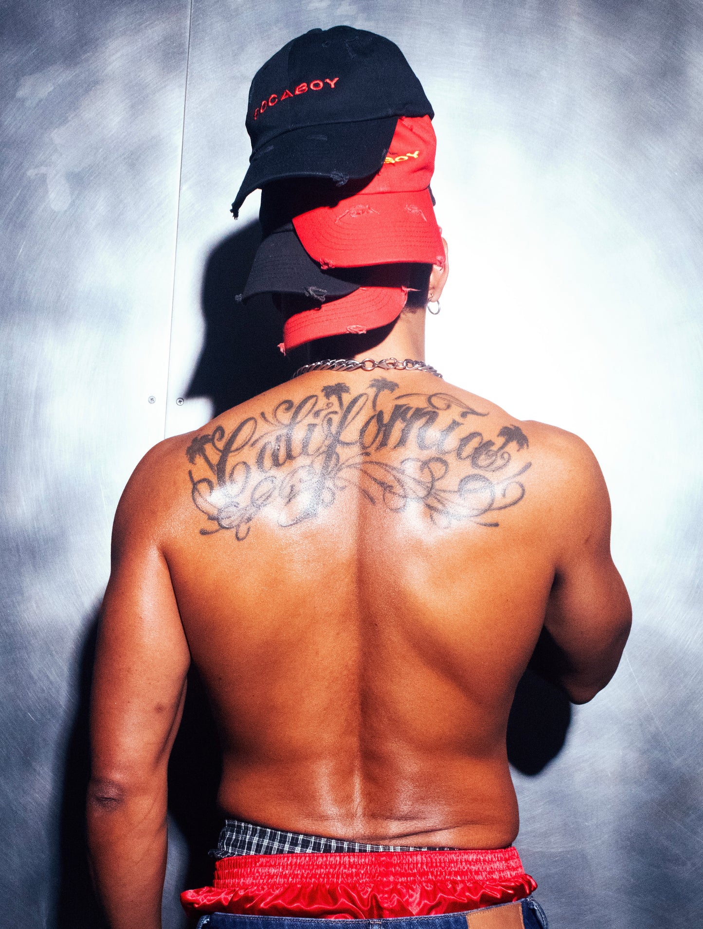 A model is photographed from the back against a brushed chrome backdrop. Four Socaboy caps in black and red are worn backwards and are stacked on top of his head. He is topless and has a 'California' tattoo across his shoulders and a pair of red satin and plaid boxer shorts are peaking over some denim jeans.