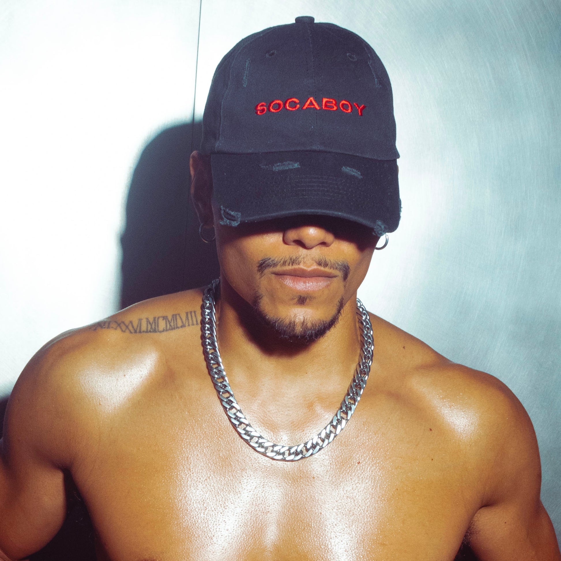 A male body wears the black Noir Cap and chunky silver chain. He is topless and features a tattoo on the left shoulder. He is standing against a brushed chrome wall.