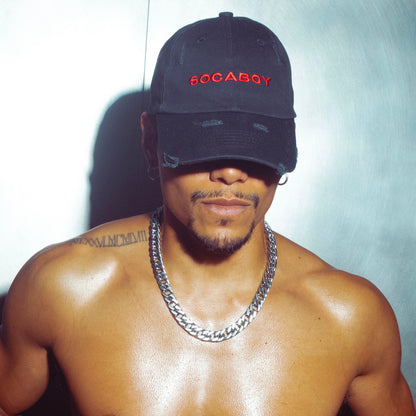 A male body wears the black Noir Cap and chunky silver chain. He is topless and features a tattoo on the left shoulder. He is standing against a brushed chrome wall.