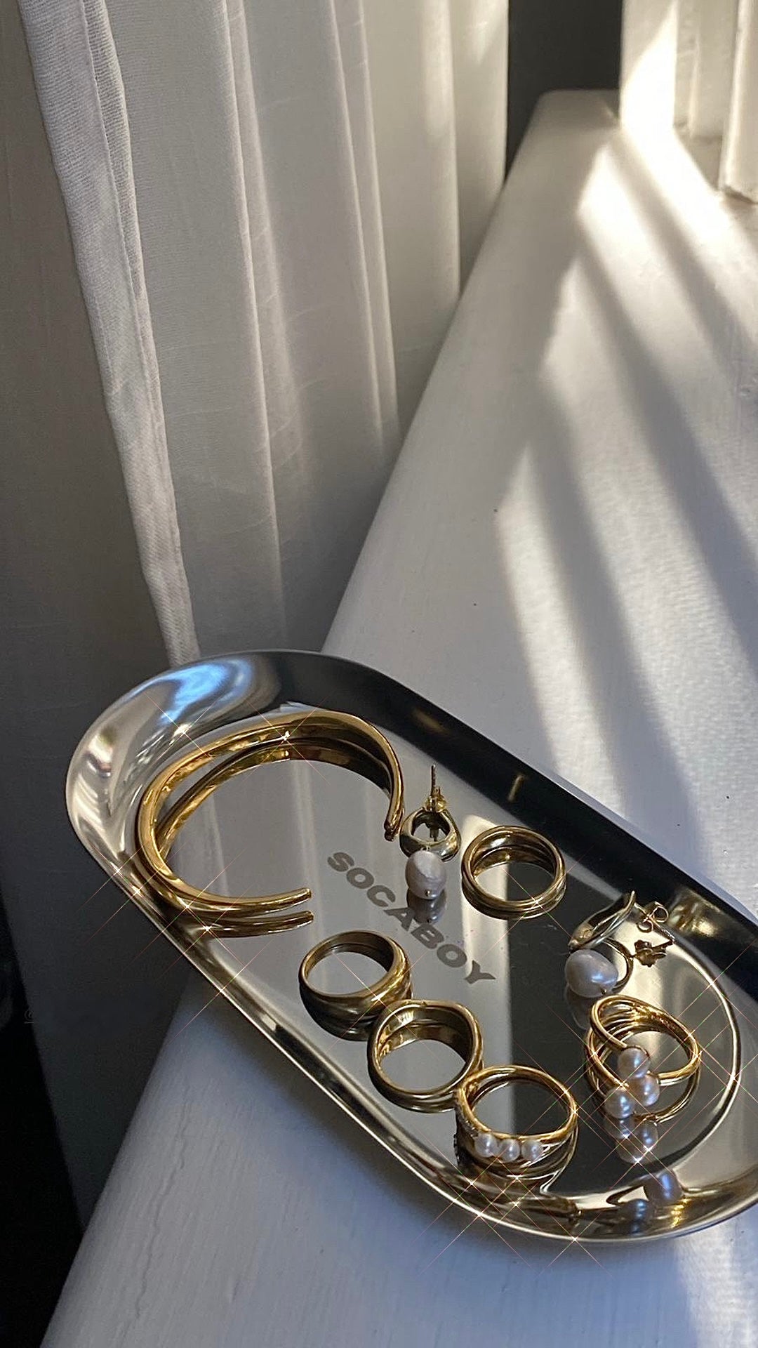Chrome Rolland Tray adorned with gold bracelet and rings with pearls, sits in the sunlight on the the window sill