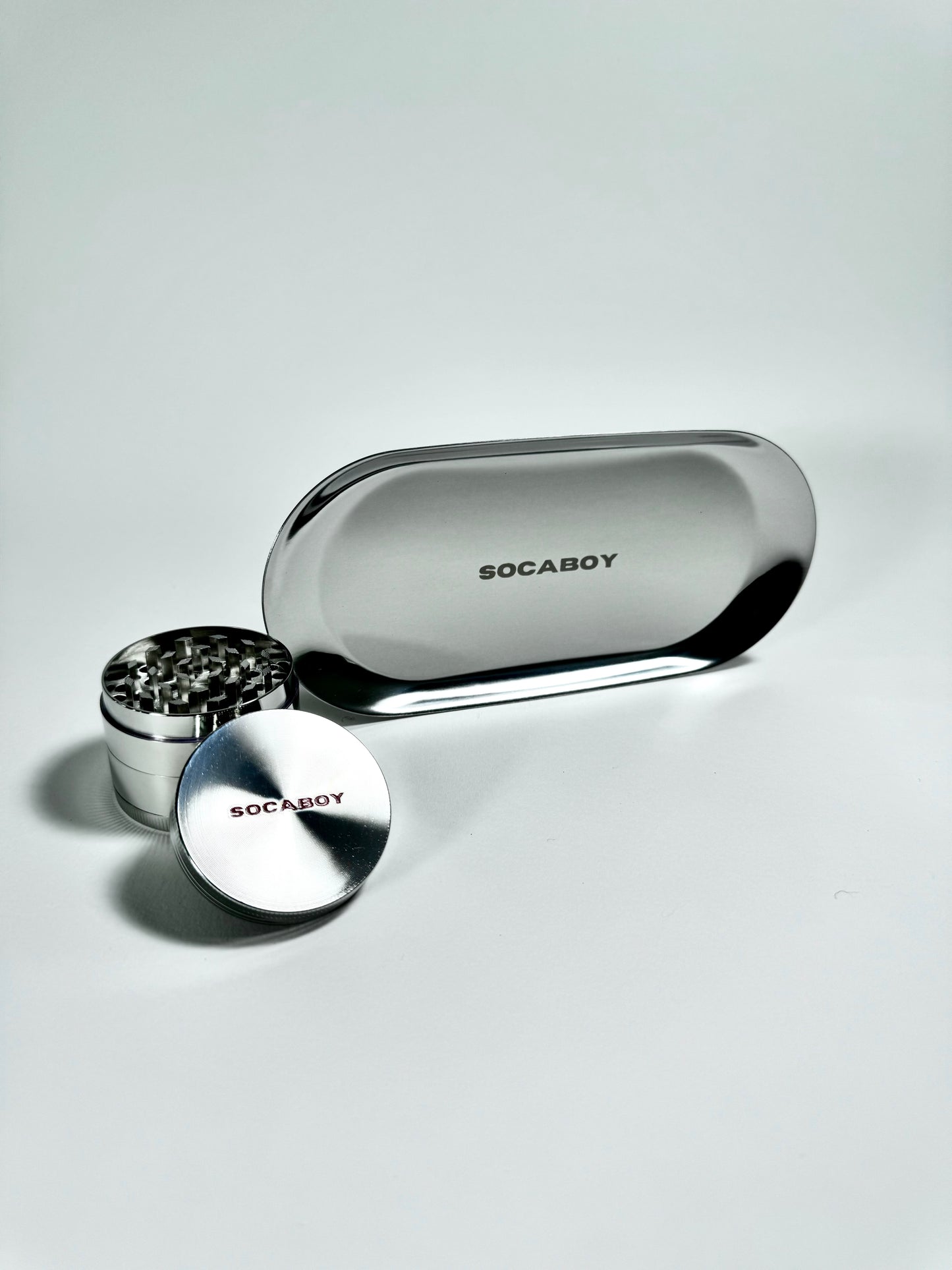 The chrome Socaboy Herb Grinder is placed in front of the chrome Rolland Tray and is photographed on a white backdrop. Both pieces feature the signature logo