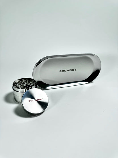 The chrome Socaboy Herb Grinder is placed in front of the chrome Rolland Tray and is photographed on a white backdrop. Both pieces feature the signature logo