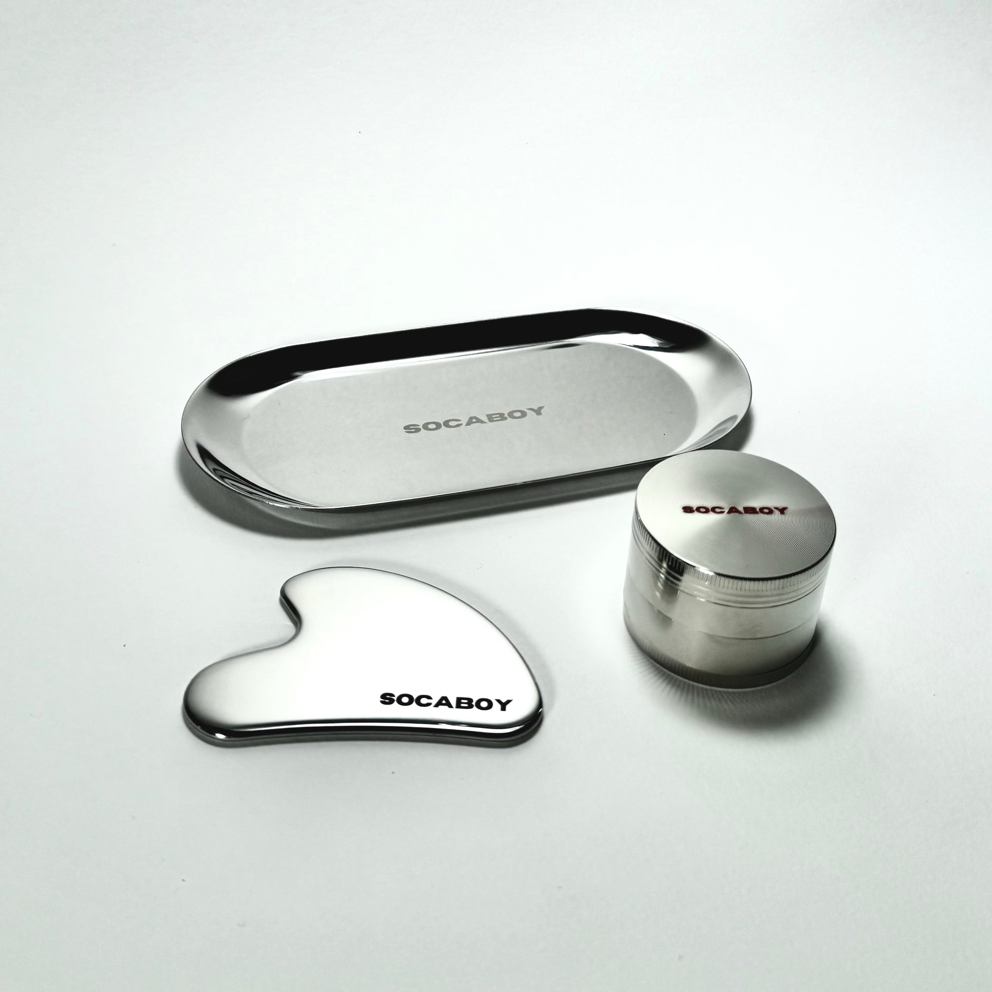 The three chrome Socaboy pieces are photographed on a white backdrop. The photo features the Rolland Tray at the top, the Gua Sha on the bototm left and the Herb Grinder on the bottom right. All three pieces feature the signature Socaboy logo.