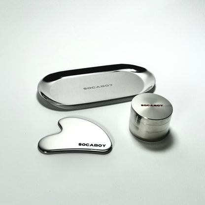 The three chrome Socaboy pieces are photographed on a white backdrop. The photo features the Rolland Tray at the top, the Gua Sha on the bototm left and the Herb Grinder on the bottom right. All three pieces feature the signature Socaboy logo.