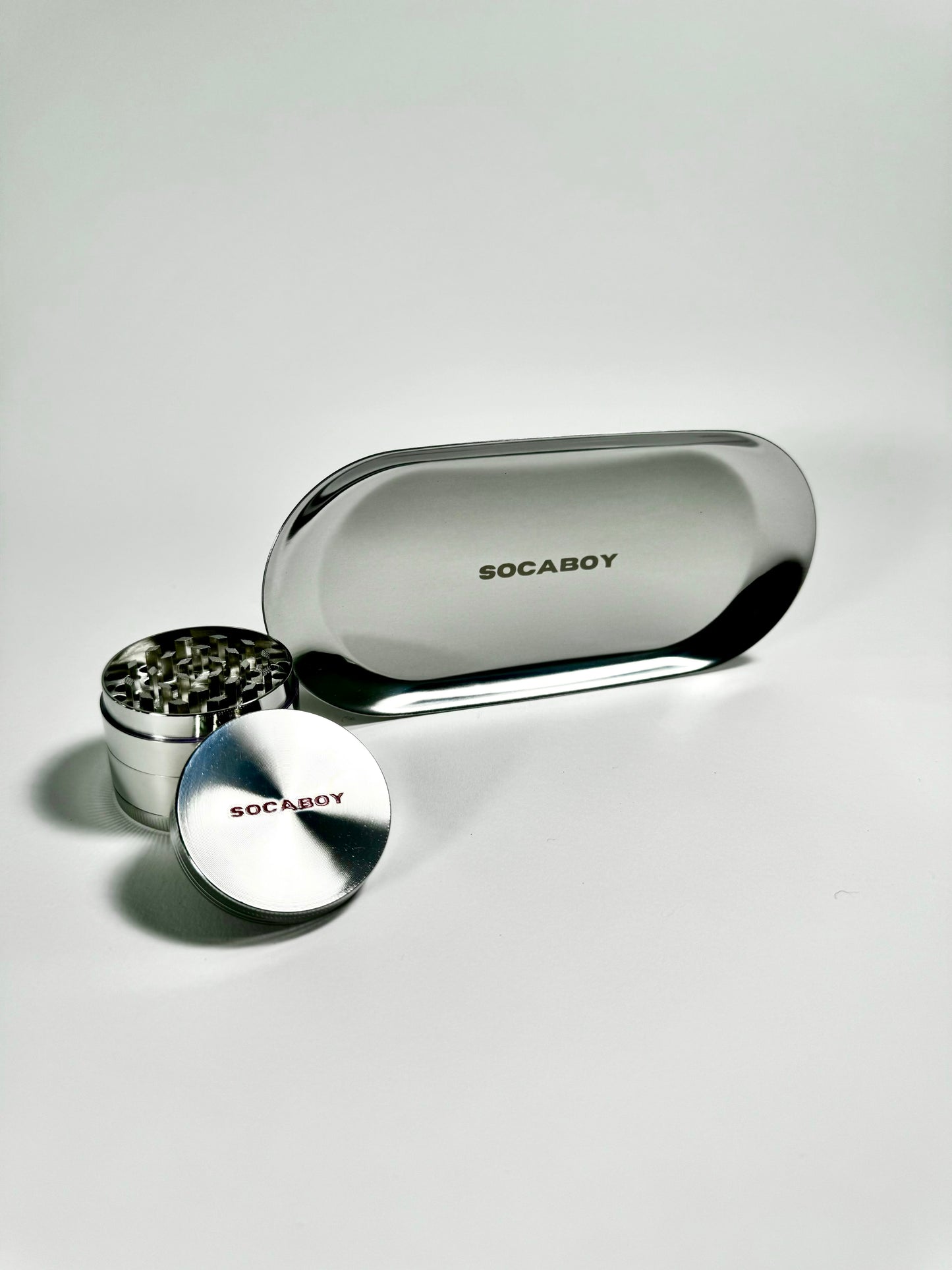 The chrome Socaboy Herb Grinder is placed in front of the chrome Rolland Tray and is photographed on a white backdrop. Both pieces feature the signature logo