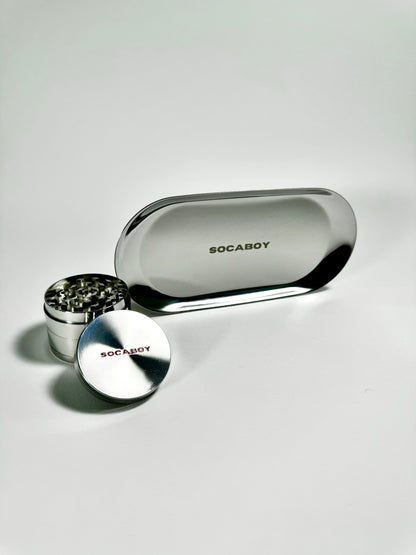 The chrome Socaboy Herb Grinder is placed in front of the chrome Rolland Tray and is photographed on a white backdrop. Both pieces feature the signature logo