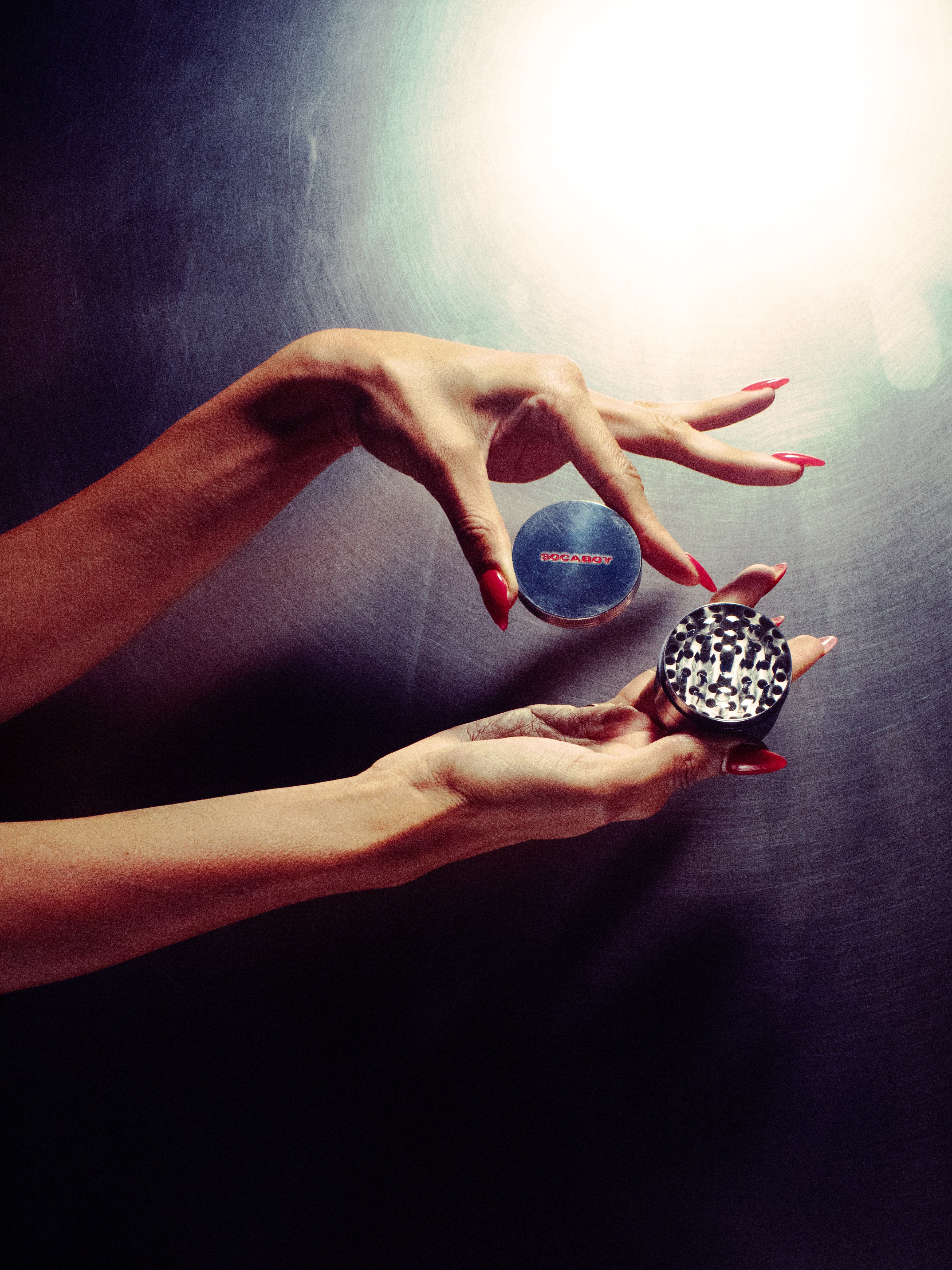 Two hands with bright red nails hold the open Socaboy Herb Grinder. The top hand holds the lid which features the logo in red. The hands and grinder are against a brushed steel backdrop