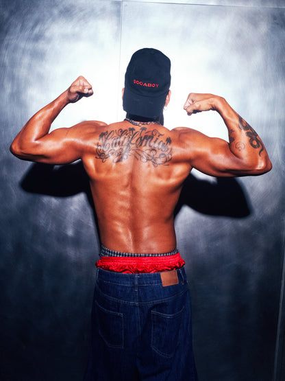 A model photographed from behind wearing the black Socaboy Noir Cap backwards. He is flexing his biceps and his muscular back reads the tattoo 'California'. He is wearing denim cut off jeans and has a pair of red satin and plaid boxer shorts peaking over the top.