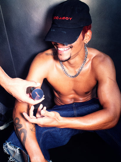 A model wearing the Socaboy Noir Cap is being handed the Socaboy Herb Grinder. He is wearing denim cut off jeans, a chunky silver necklace and is smiling.