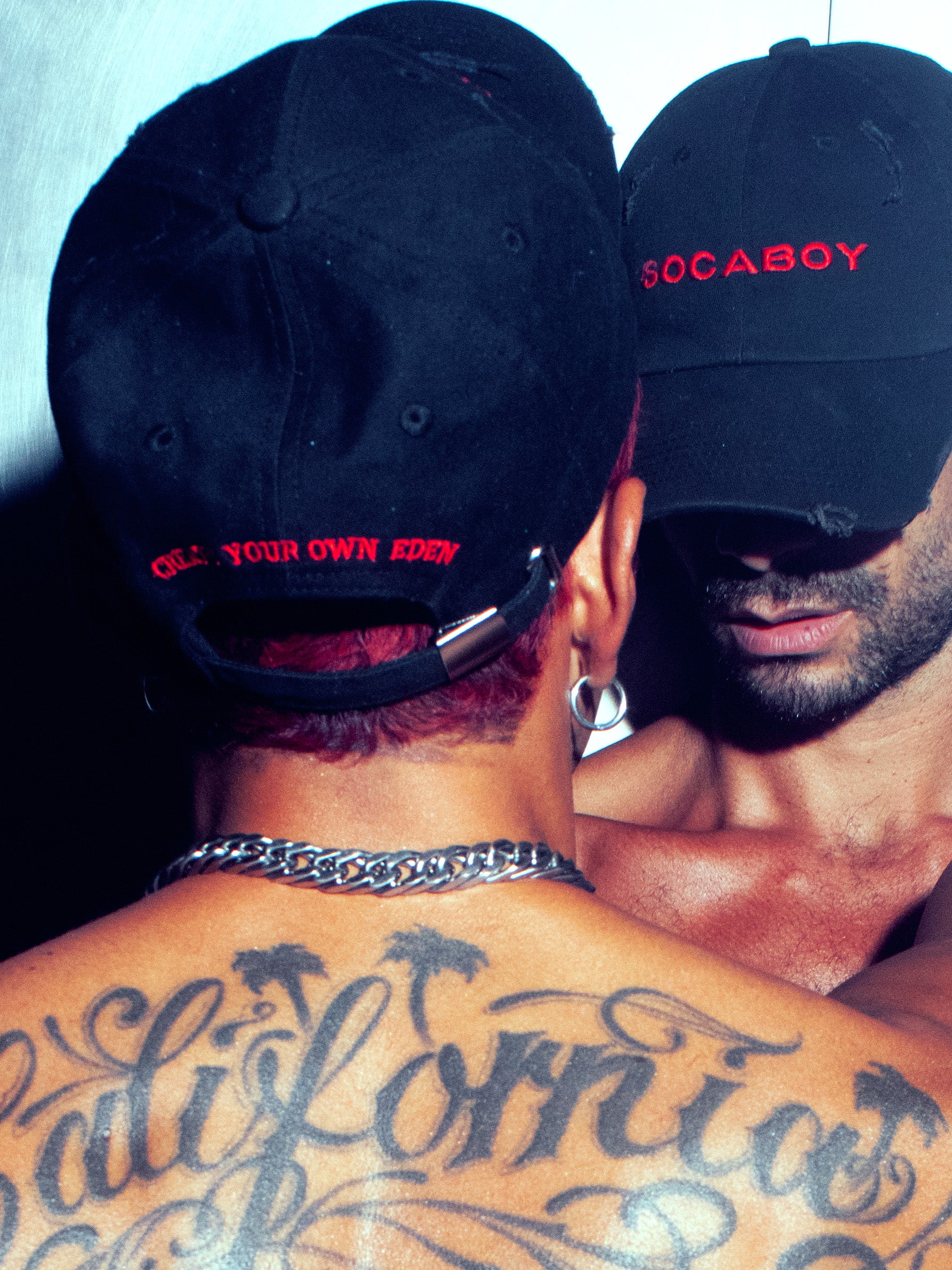 Two shirtless models facing each other wearing the Socaboy Noir Caps. The back of one model is in the foreground featuring the cap with the embroidered affirmation 'create your own eden'; he is wearing a chunky silver necklace and silver hoop earrings. The other model is facing him with his eyes covered by the cap. 