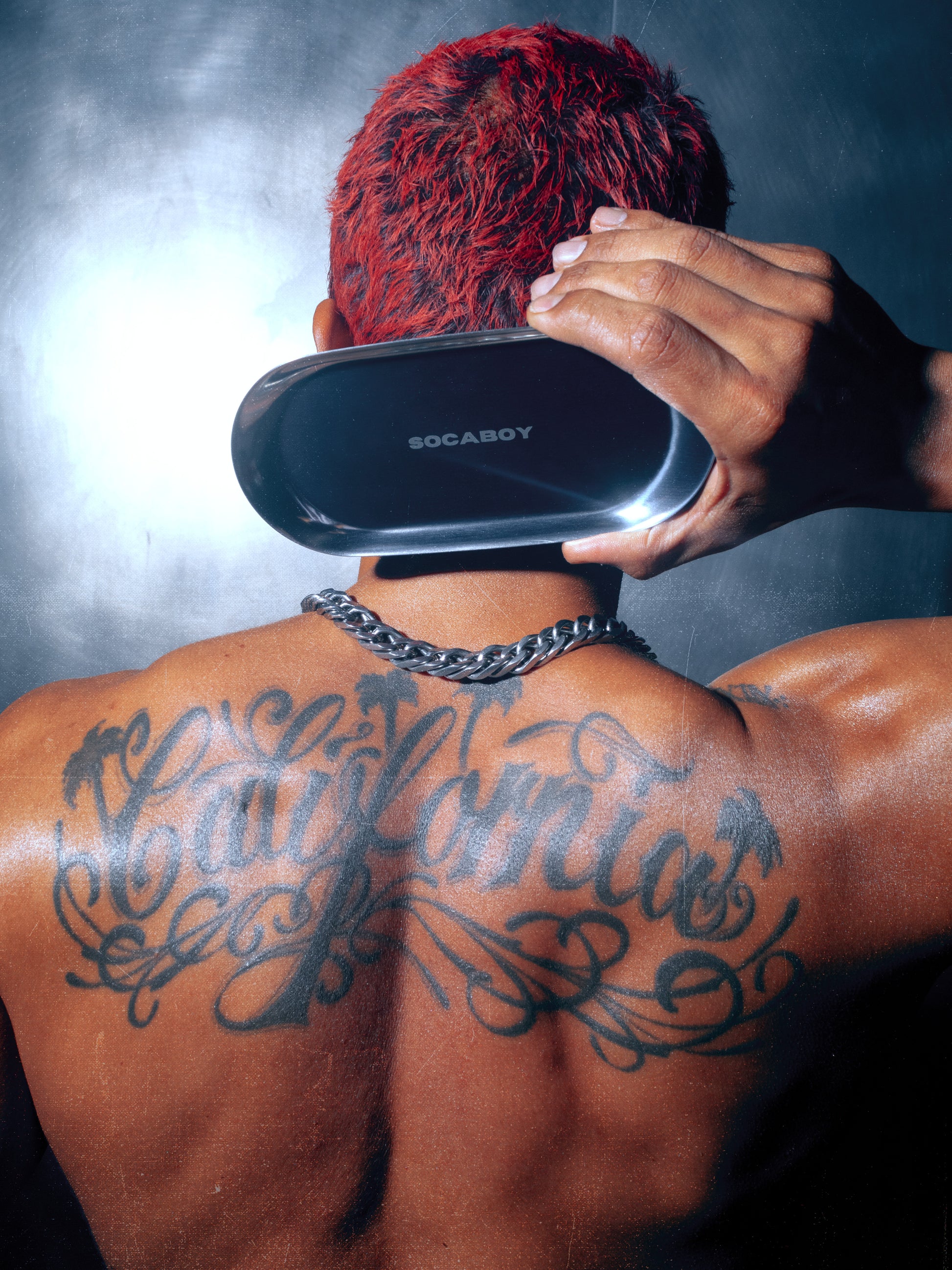 A model holds the Socaboy Rolland Tray in his right hand behind the back of his head against a brushed chrome wall.  He has a muscular back with a large cursive tattoo across his shoulders of the word 'California' He has flame red hair and a chunky silver chain arond his neck. The Rolland Tray shines in the light.