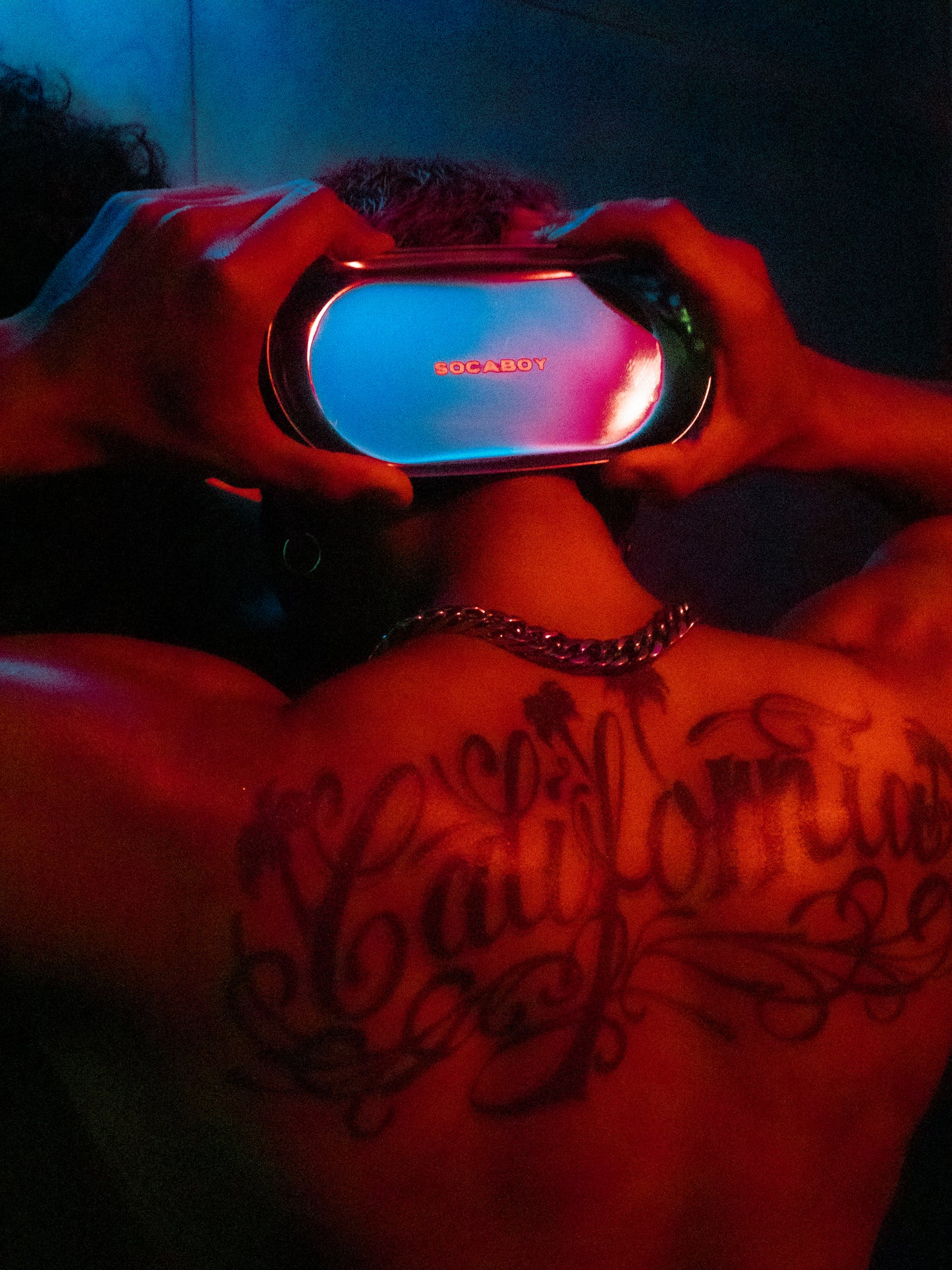 A model holds the Rolland Tray behind his head with both hands. He is covered in a red light. His back features a large tattoo of the word 'California' and his wearing a chunky silver chain. 