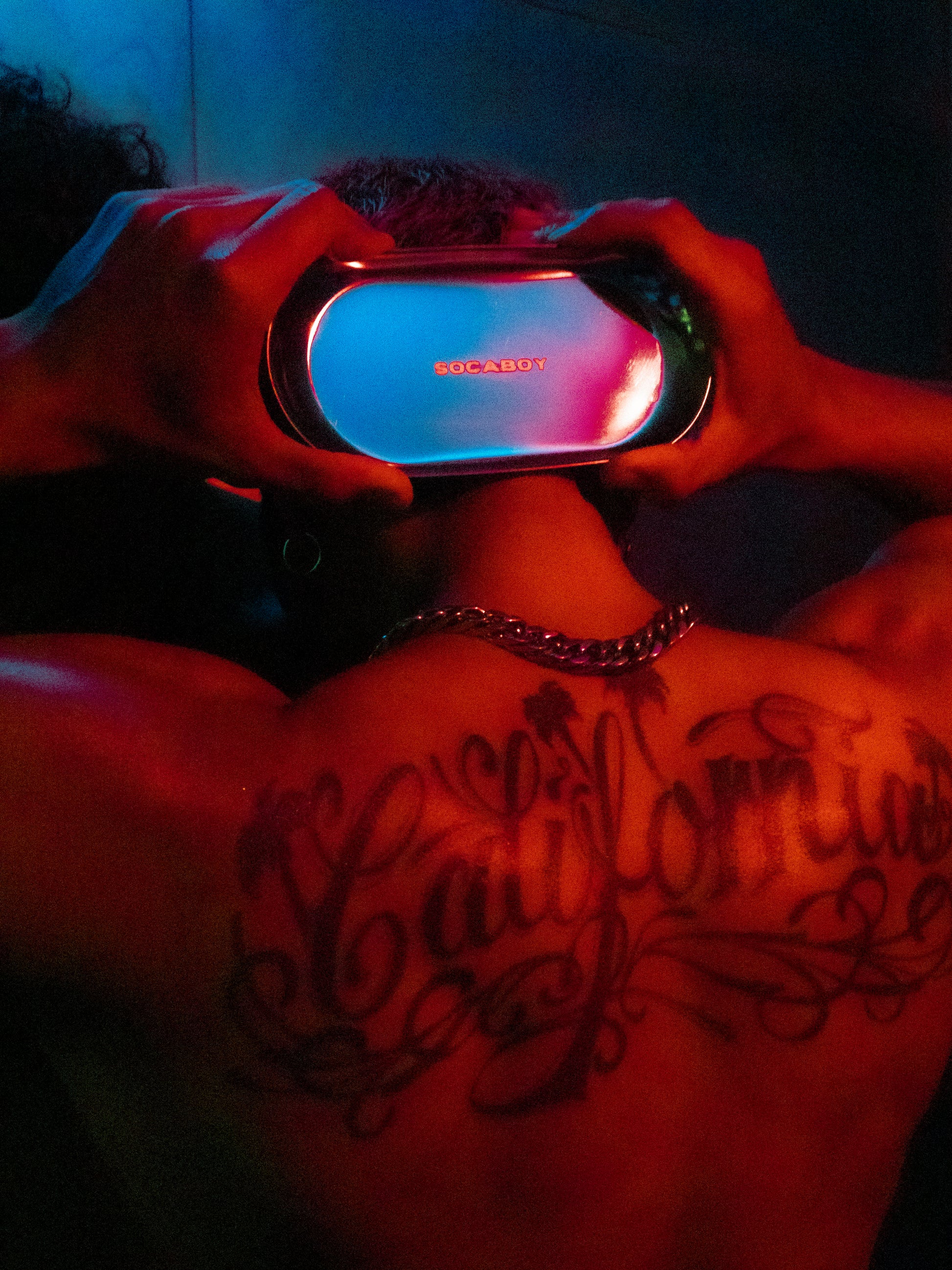 A model holds the Rolland Tray behind his head with both hands. He is covered in a red light. His back features a large tattoo of the word 'California' and his wearing a chunky silver chain. 