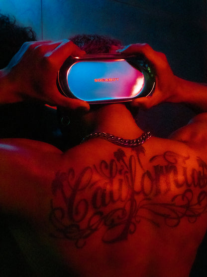 A model holds the Rolland Tray behind his head with both hands. He is covered in a red light. His back features a large tattoo of the word 'California' and his wearing a chunky silver chain. 