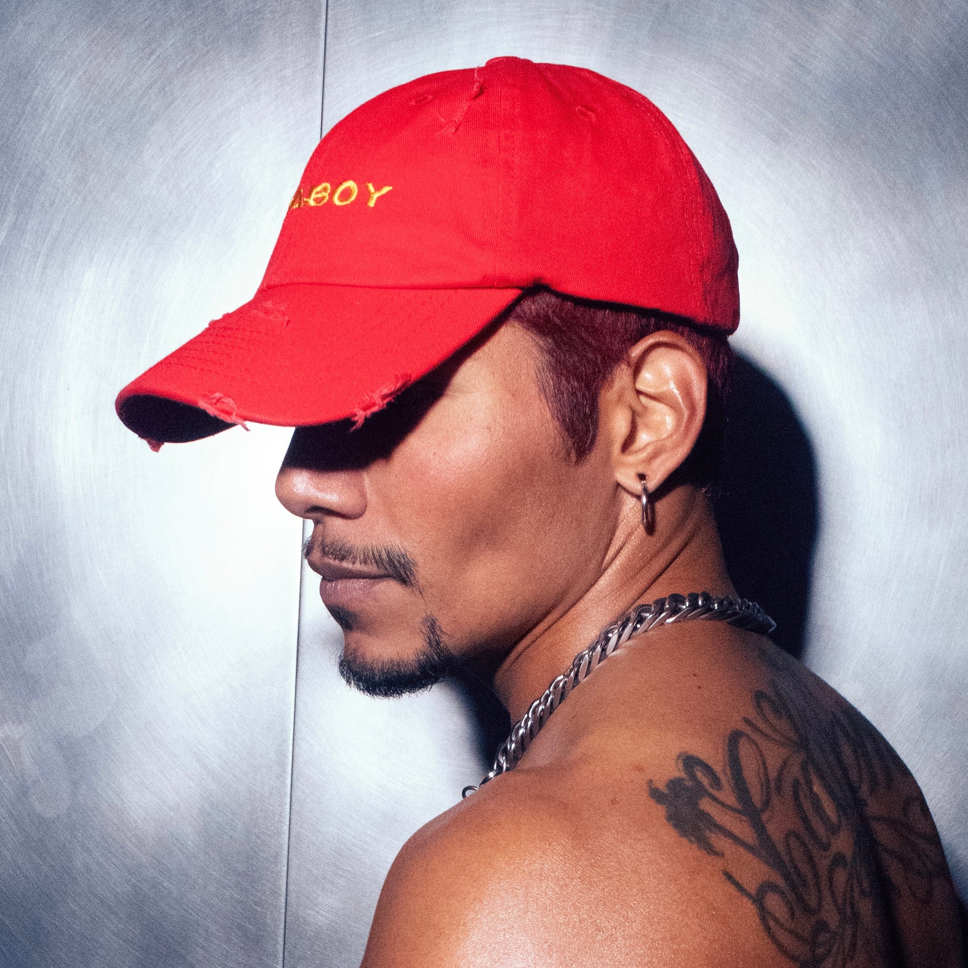 A model is standing profile against a chrome wall wearing the red Socaboy Rossa Cap. The cap covers his eyes and he is shirtless with a chunky silver necklace and back tattoo.