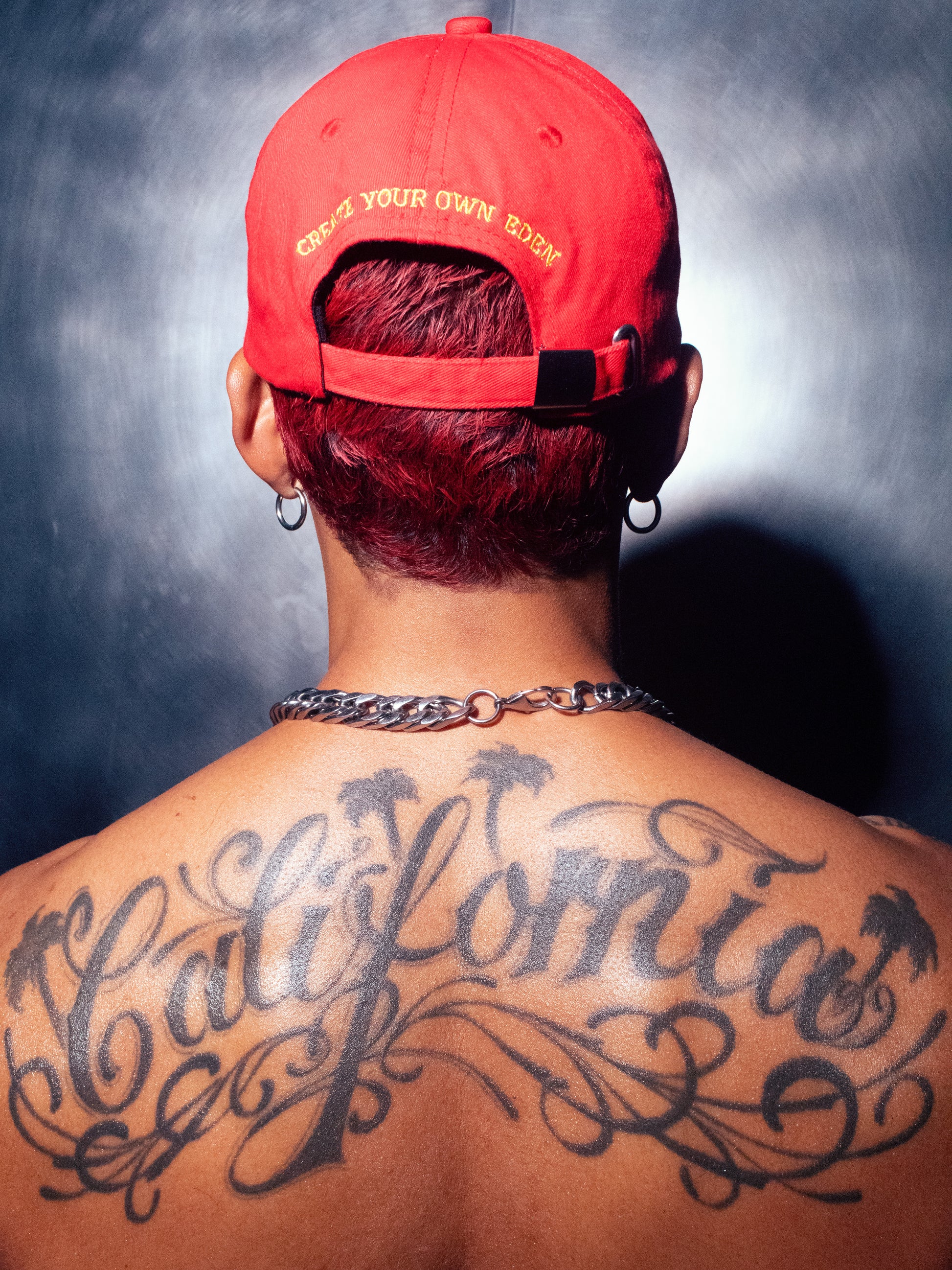 A photograph of the back of a model wearing the red Socaboy Rossa Cap which features the affirmation 'create your own eden' in yellow. The model is shirtless and is wearing a chunky silver chain and two silver earrings and has a large back tattoo of the word "California'. He is standing against a brushed chrome wall.