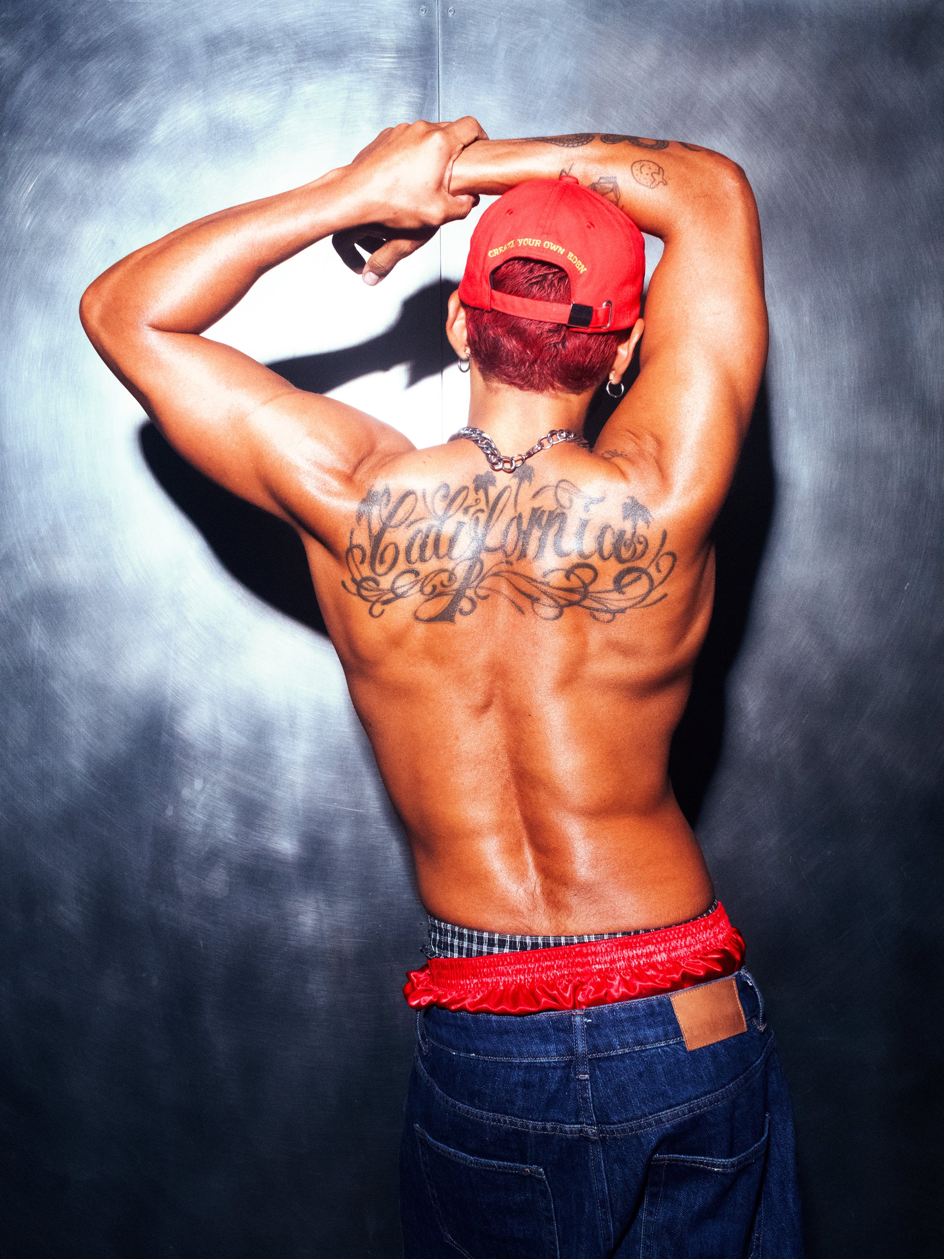 A model photographed from behind wearing the red Socaboy Noir Cap backwards. He has his hands over his head and his muscular back reads the tattoo 'California'. He is wearing denim cut off jeans and has a pair of red satin and plaid boxer shorts peaking over the top.