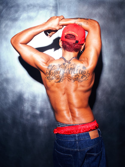 A model photographed from behind wearing the red Socaboy Noir Cap backwards. He has his hands over his head and his muscular back reads the tattoo 'California'. He is wearing denim cut off jeans and has a pair of red satin and plaid boxer shorts peaking over the top.