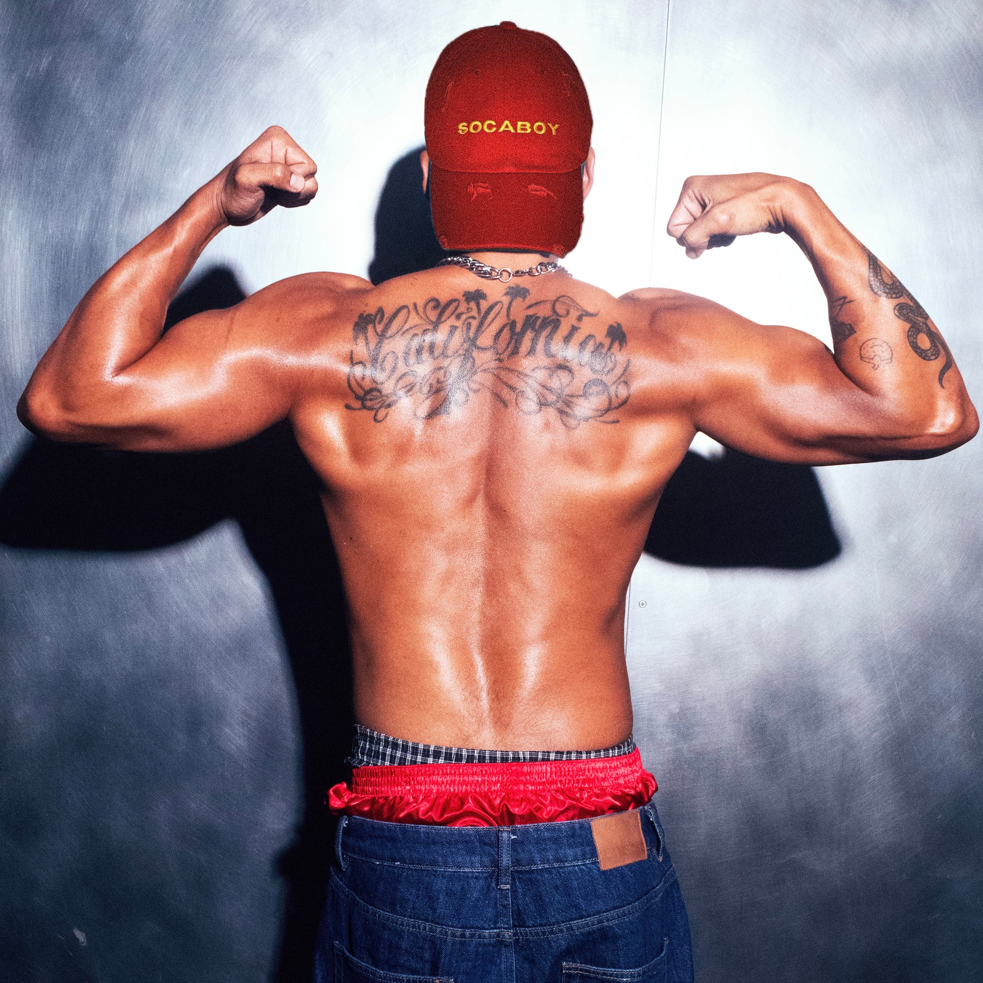 A model photographed from behind wearing the red Socaboy Rossa Cap backwards. He is flexing his biceps and his muscular back reads the tattoo 'California'. He is wearing denim cut off jeans and has a pair of red satin and plaid boxer shorts peaking over the top.