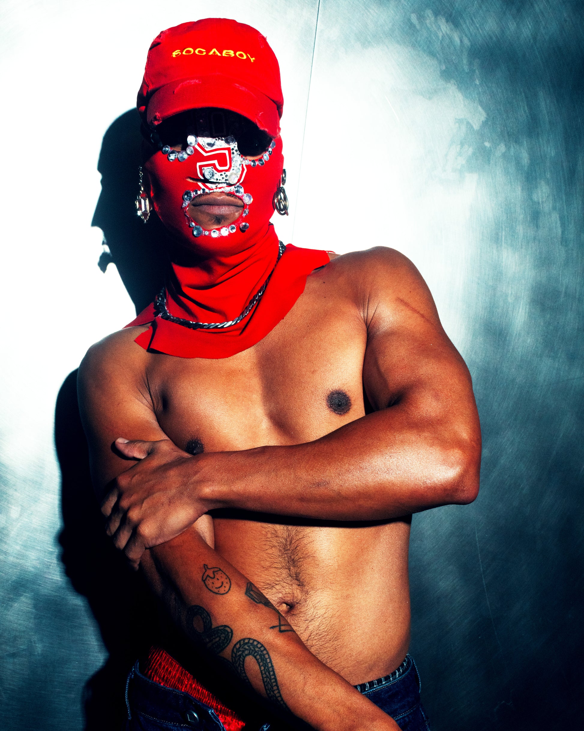 A model wearing a custom red jewelled ski mask and the red Socaboy Rossa Cap. He is shirtless and has tattoos on his forearm.