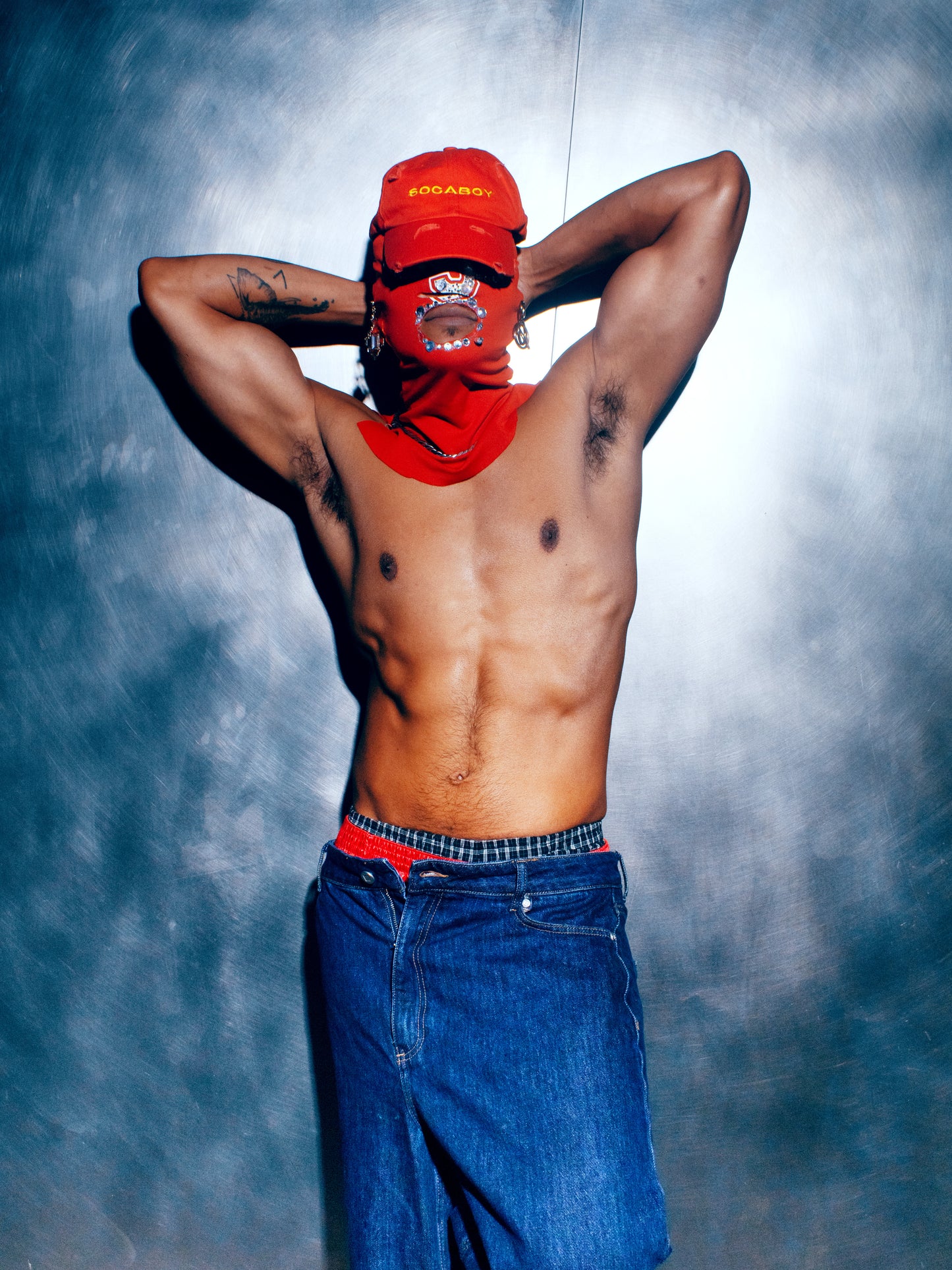 A model wearing a custom red jewelled ski mask and the red Socaboy Rossa Cap. He is shirtless, wearing denim cut off jeans and boxer shorts  and has his hands behind his head.